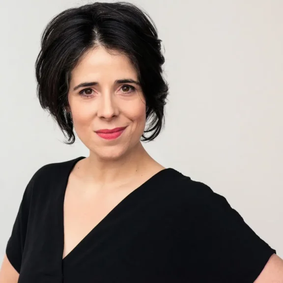 Woman with short dark hair, wearing a black V-neck top, looks at the camera with a slight smile against a plain light background.
