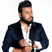 A man with dark hair and beard wearing a patterned navy suit and white shirt adjusts his cuff while looking to the side against a plain white background.