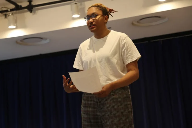 A person wearing a white t-shirt and plaid pants stands on a stage, holding a sheet of paper and speaking.