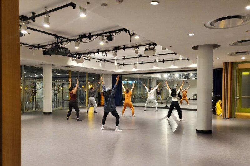 A group of people in workout attire practice dance or exercise movements in a brightly lit studio with large windows.