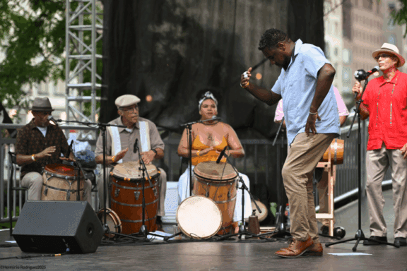 A man dances onstage in front of a seated musical group playing drums and percussion instruments during an outdoor performance.