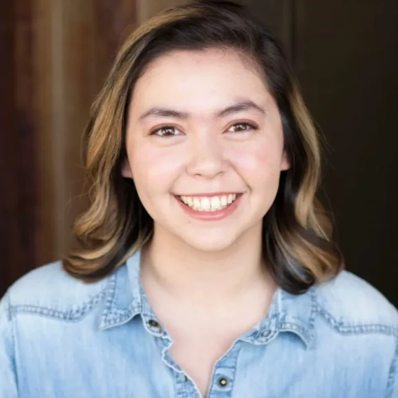 A young woman with shoulder-length light brown hair smiles at the camera. She is wearing a light blue denim shirt and is standing in front of a blurred indoor background.