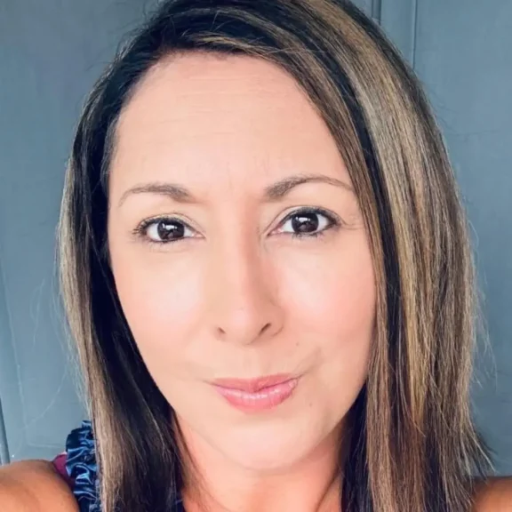 A woman with straight, shoulder-length brown hair and light highlights looks directly at the camera against a plain background.