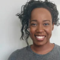 A woman with curly hair smiles at the camera, wearing a grey shirt against a plain light background.