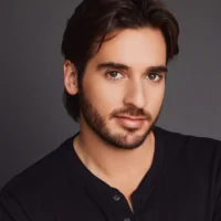 A man with medium-length dark hair, a trimmed beard, and mustache, wearing a black buttoned shirt, poses in front of a plain dark background.