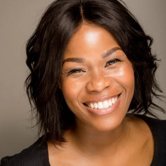 Woman with shoulder-length dark hair smiles broadly, showing teeth, against a neutral background. She is wearing a black top.