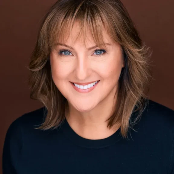 Woman with light brown hair and bangs, wearing a black top, smiling at the camera against a plain brown background.