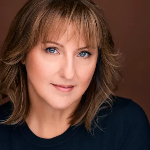 A woman with light brown hair and blue eyes poses in front of a brown background, wearing a dark top and a neutral expression.