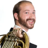 A man with a beard and short hair, wearing a tuxedo, smiles at the camera while holding a French horn over his shoulder against a white background.