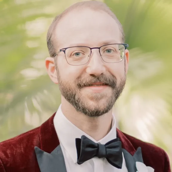 A man with glasses, a beard, and a mustache wears a burgundy velvet tuxedo jacket, white shirt, and black bow tie, standing in front of a blurred green background.