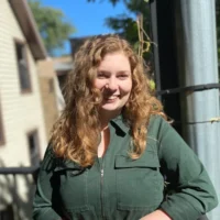 A woman with curly light brown hair, wearing a green zip-up shirt, stands outside in sunlight, smiling at the camera.