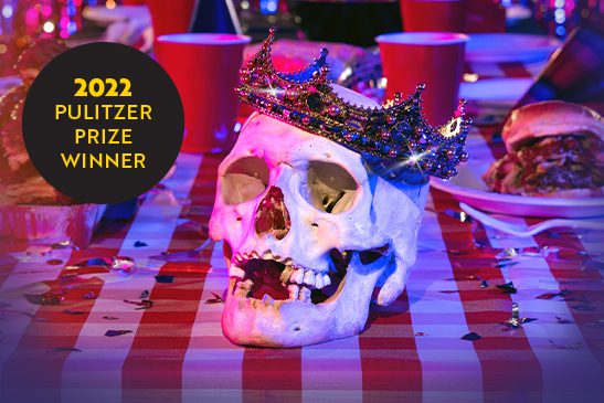 A jewel-encrusted crown adorns a skull sitting on a table littered with red Solo cups and barbecued meats.