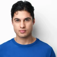 A young man with short dark hair and a neutral expression, wearing a blue shirt, stands in front of a plain white background.