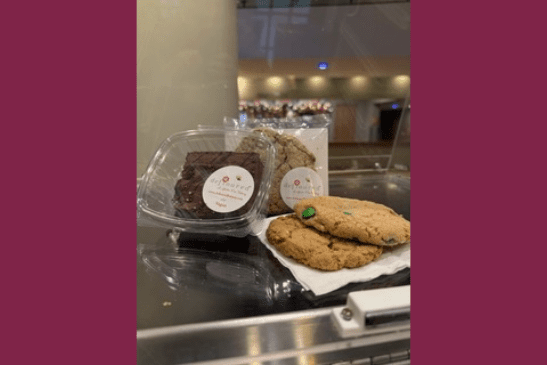 A brownie in a to-go box, a wrapped chocolate-chip cookie, and two m&m's cookies on a napkin.