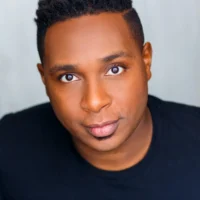 A person with short, curly black hair and brown eyes wears a black shirt and looks directly at the camera against a plain background.
