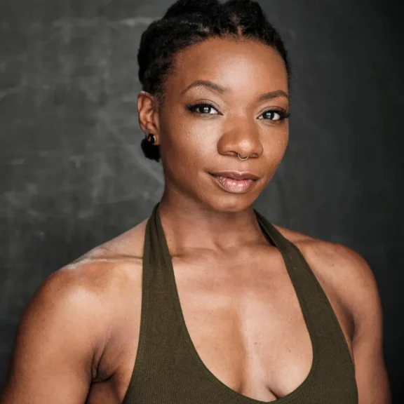 A woman with short, braided hair and a nose ring poses against a dark background, wearing a sleeveless olive green top.