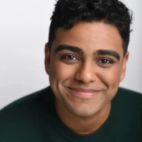A young person with curly dark hair and medium skin tone smiles softly, wearing a dark green top, against a plain light background.