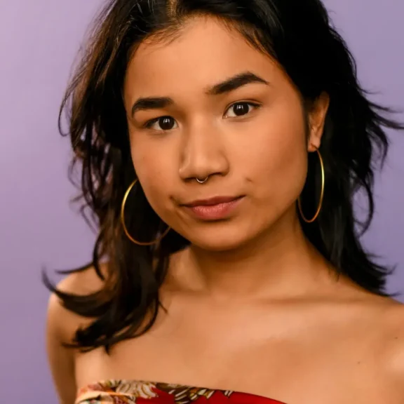 Person with medium-length dark hair, gold hoop earrings, and a nose ring, wearing a red, off-the-shoulder top, poses in front of a plain purple background.