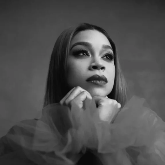 A woman with long hair gazes upward with her chin resting on her hands, wearing a sheer, tulle-like garment in a black-and-white portrait.