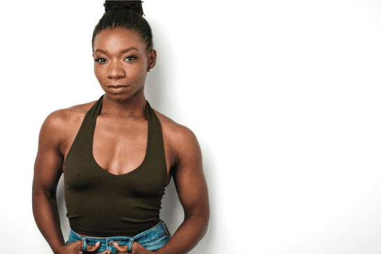 A black person with hair in a bun, an olive green halter tank top, and blue jeans.