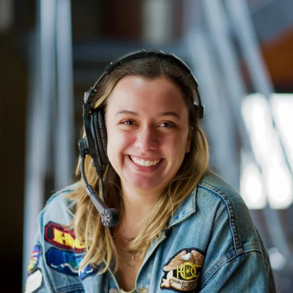 A person wearing a headset and a denim jacket with patches smiles at the camera in an indoor setting.