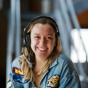 Person wears headset, has blonde hair, and wears denim jacket with patches.