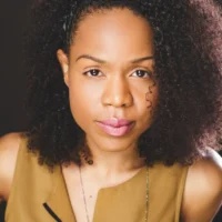 A woman with curly hair and a neutral expression looks directly at the camera. She is wearing a tan sleeveless top and a necklace. The background is dark.