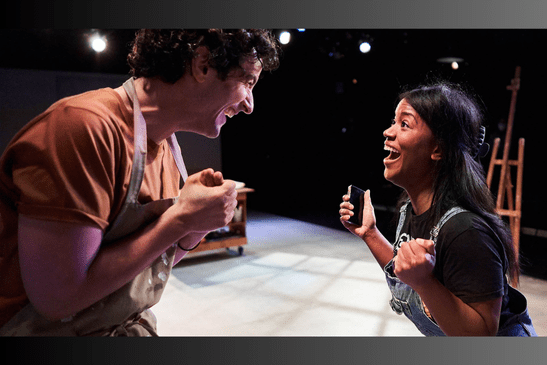 Two actors stand face-to-face on the theater set of an artist's studio. They are both wearing smocks and smiling excitedly at one another.