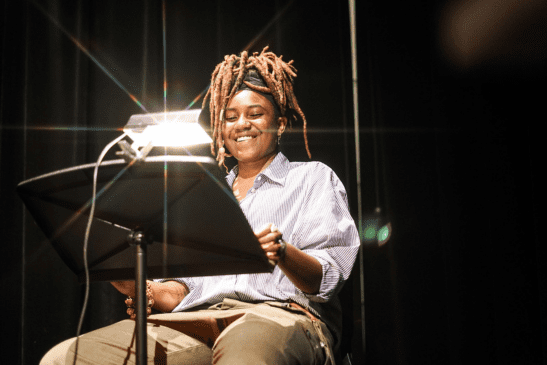 A smiling actor reads from a script on a music stand, illuminated by lamplight.