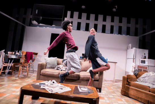 Two actors stand face-to-face on the theater set of an artist's studio. They are both wearing smocks and smiling excitedly at one another.