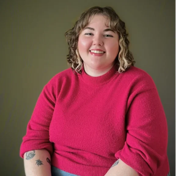 A person with curly hair and tattoos on their forearms sits smiling, wearing a bright pink sweater against a plain, neutral background.
