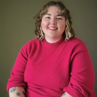 A person with curly hair and tattoos on their arms is smiling while wearing a bright pink sweater, sitting against a plain background.