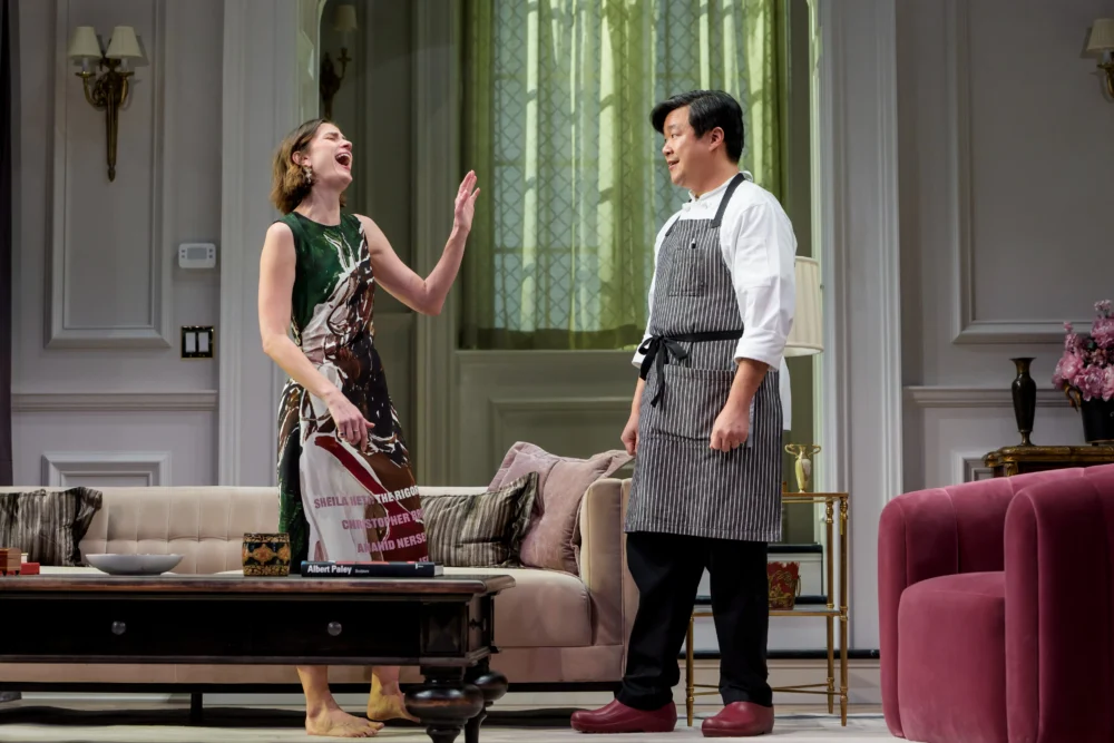 A woman in a dress laughs and raises her hand while talking to a man in a chef’s uniform in a modern, elegantly decorated living room.