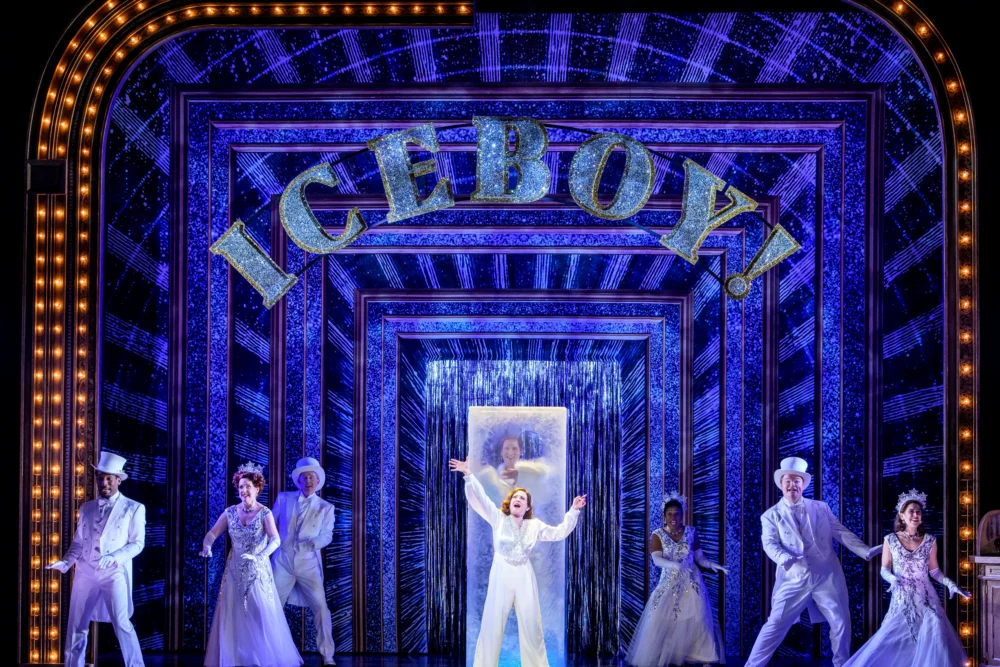 Performers in white costumes dance on stage in front of a sparkling set with the word "ICEBOY!" lit up above, framed by bright marquee lights.