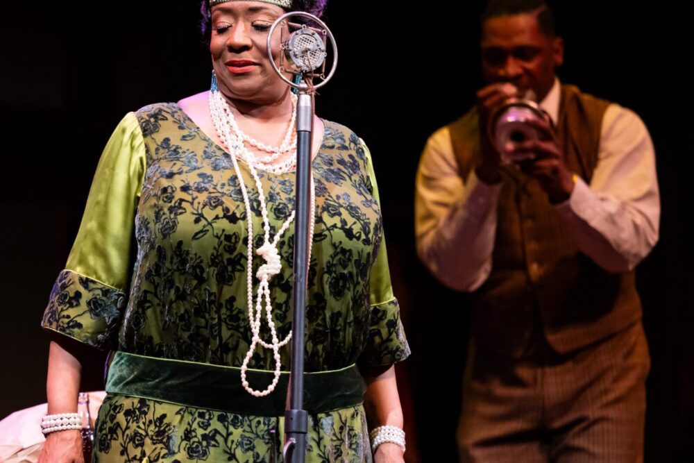 A woman in a green and black dress stands at a vintage microphone while a man in the background plays a trumpet.