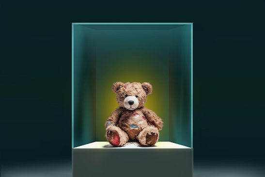 A teddy bear, its fur patchy from years of affection, sits inside a brightly lit glass vitrine as if on display at a museum.