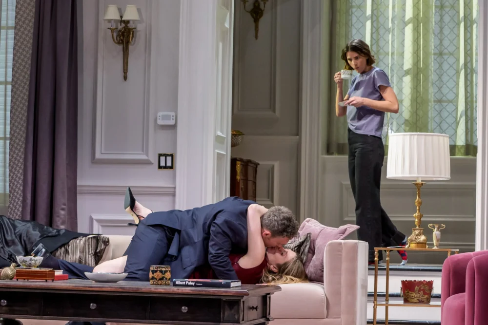 A man and woman kiss passionately on a couch while another woman stands in the background watching them from a distance.