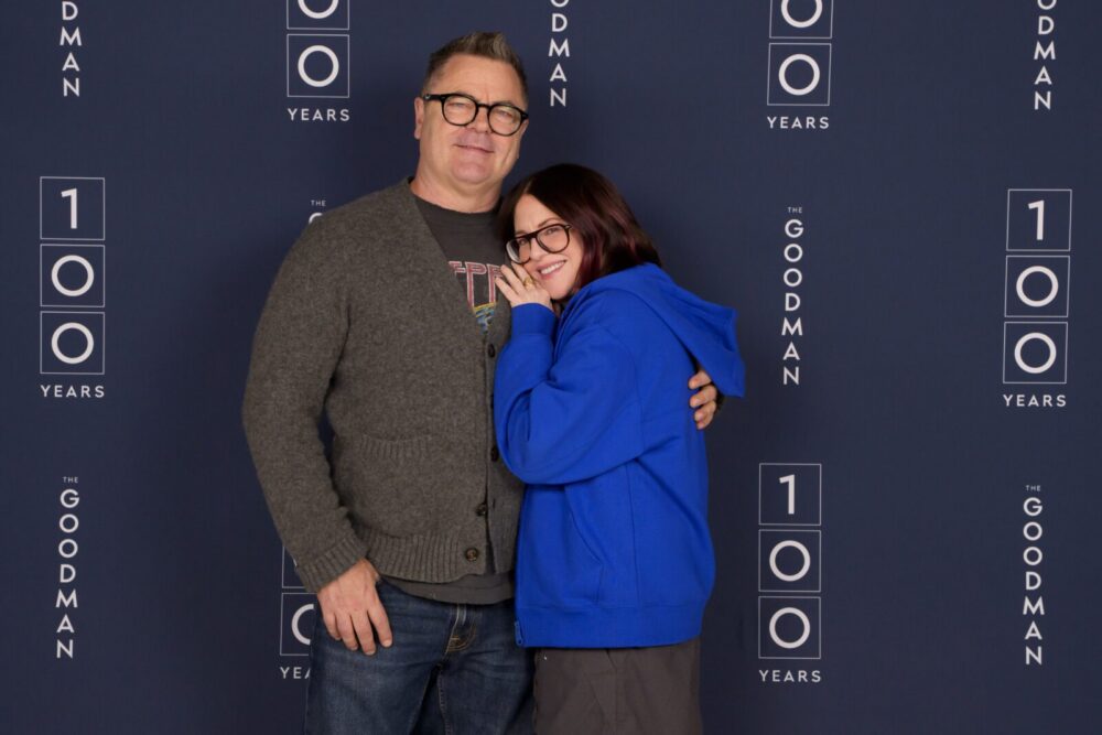 Two people pose together in front of a blue backdrop with "100 Years" and "The Goodman" printed repeatedly in white text.