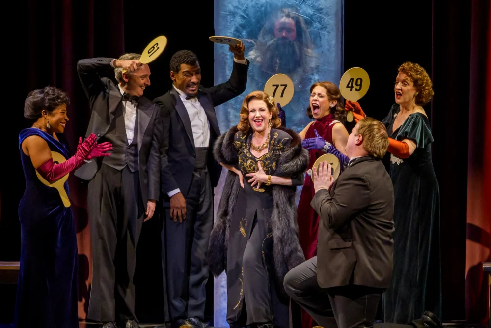 Seven people in formal attire hold numbered paddles and gesture animatedly on stage; a bearded man is visible behind them, encased in a large clear box.