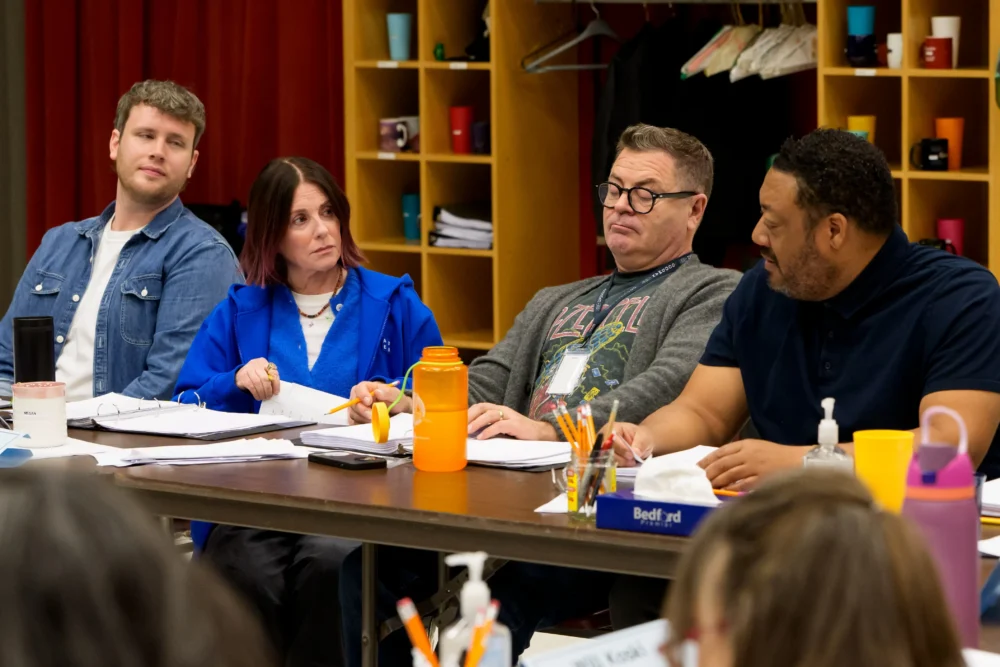 Four people sit at a table with notebooks, pens, and drinks, engaged in conversation during a meeting in a brightly lit room with shelves in the background.