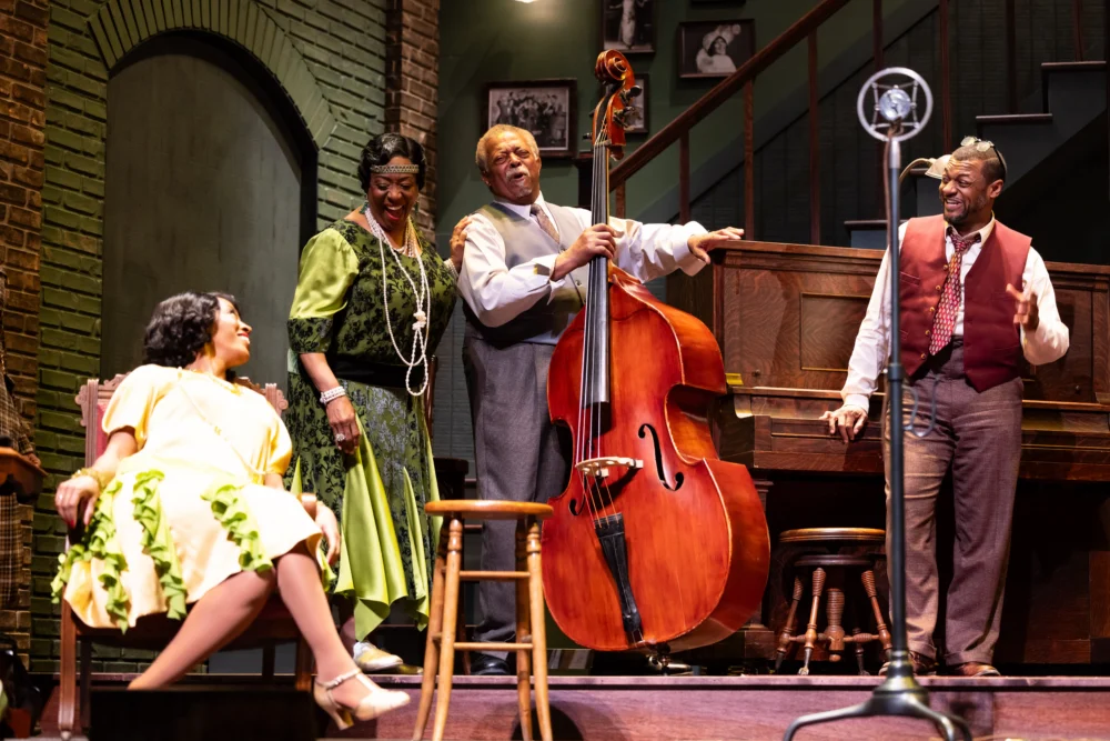 Four performers in period costumes are on stage; one sits, two stand near a double bass, and one stands near a piano and vintage microphone, all appearing engaged and expressive.