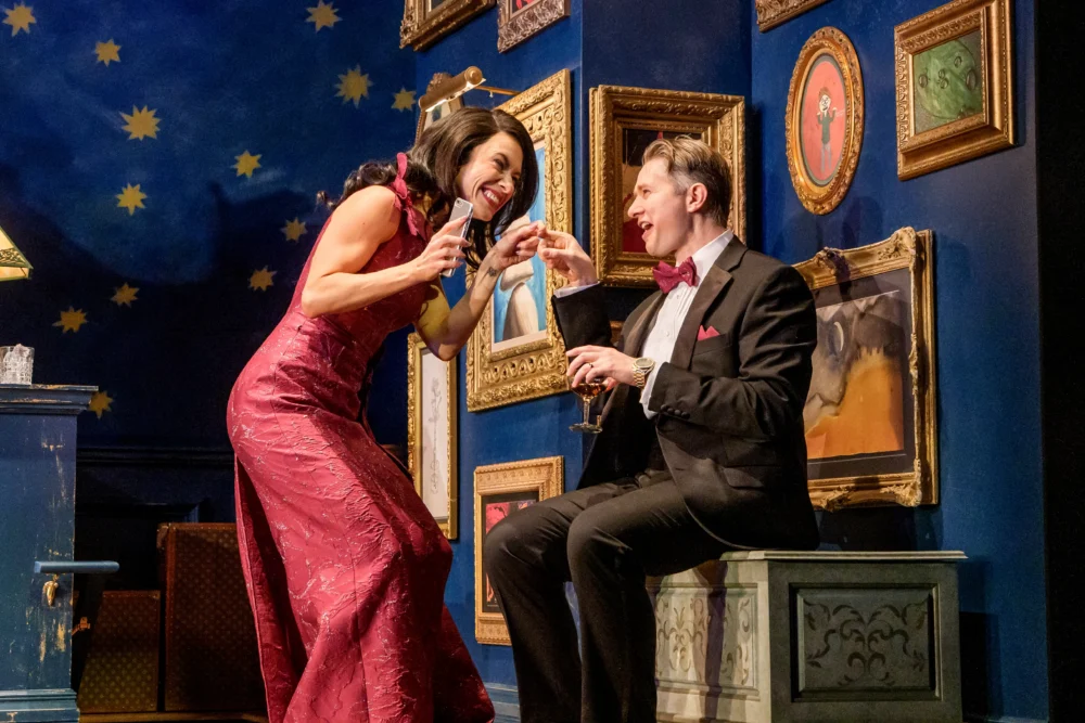 A woman in a red dress and a man in a tuxedo smile and toast with wine glasses in a brightly decorated room with framed art and a starry sky backdrop.