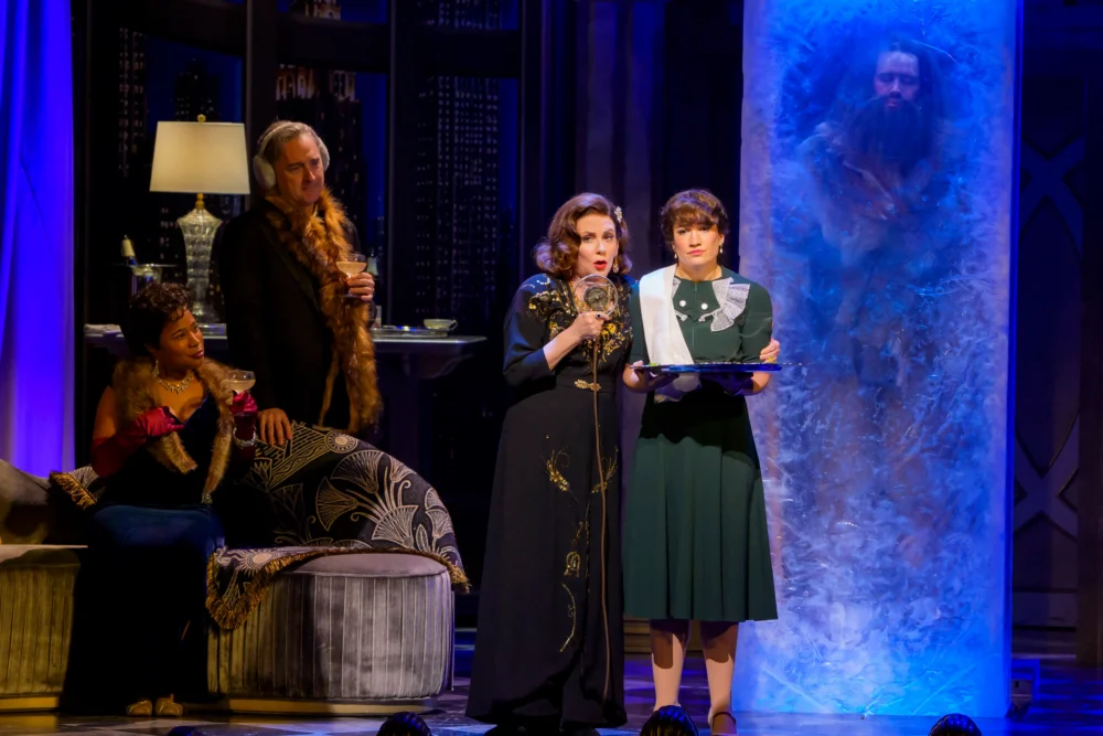 Four people are onstage in vintage attire; one man is frozen inside a large upright block of ice, while others watch, one woman holding a tray. The set resembles a lavish living room.