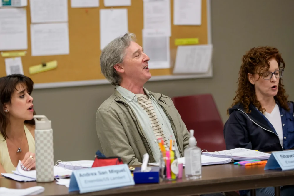 Three people sit at a table in a meeting room, appearing to sing or speak together. Papers, pens, and water bottles are on the table, with a bulletin board in the background.
