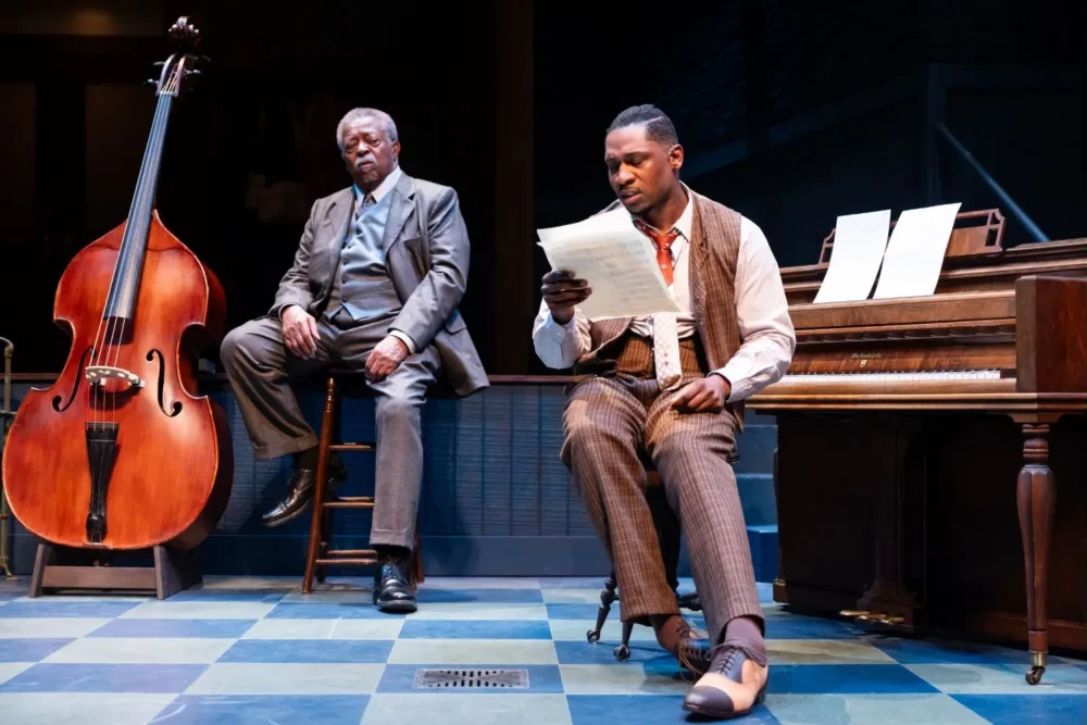 Two men in suits are on stage; one sits on a stool beside a double bass, the other sits at a piano reading sheet music.