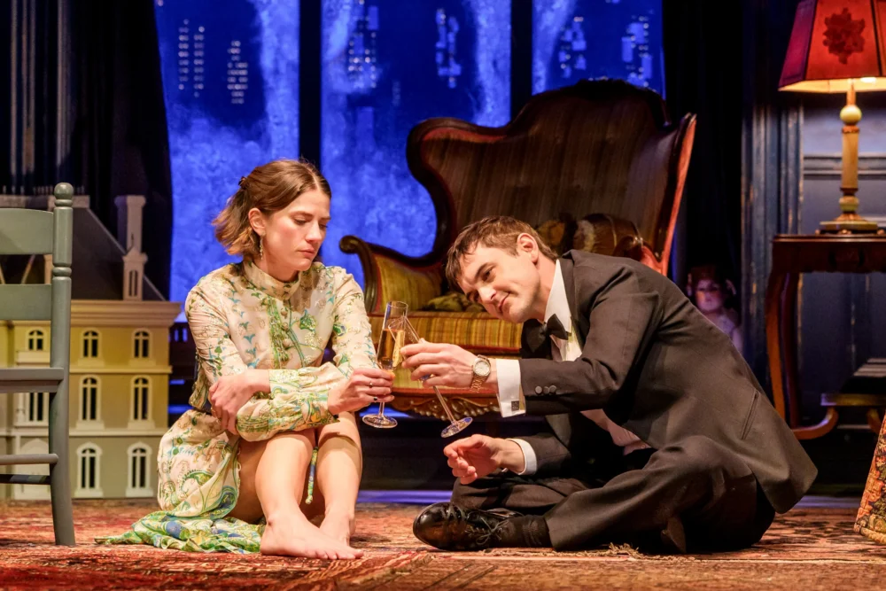 A woman in a floral dress and a man in a suit sit on a rug, clinking champagne glasses, with a dollhouse and vintage furniture in the background.