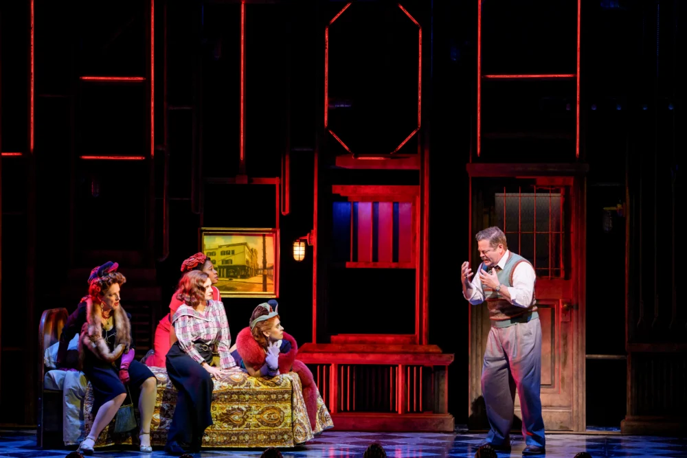 Five actors perform a dramatic scene on stage, with one man standing and gesturing while four people sit on a couch, set against a backdrop with red lighting and industrial elements.