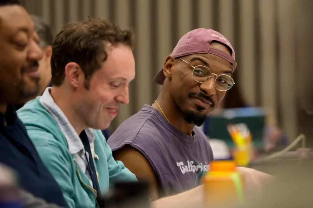 Three people sit side by side at a table in conversation; one man in glasses and a backward cap looks toward the camera.
