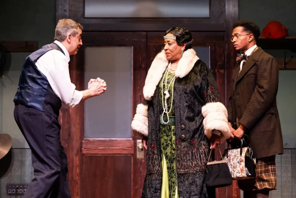 Three actors perform on stage; one man gestures with a handkerchief, while a woman in a fur-trimmed coat and another man stand facing him near a wooden door.