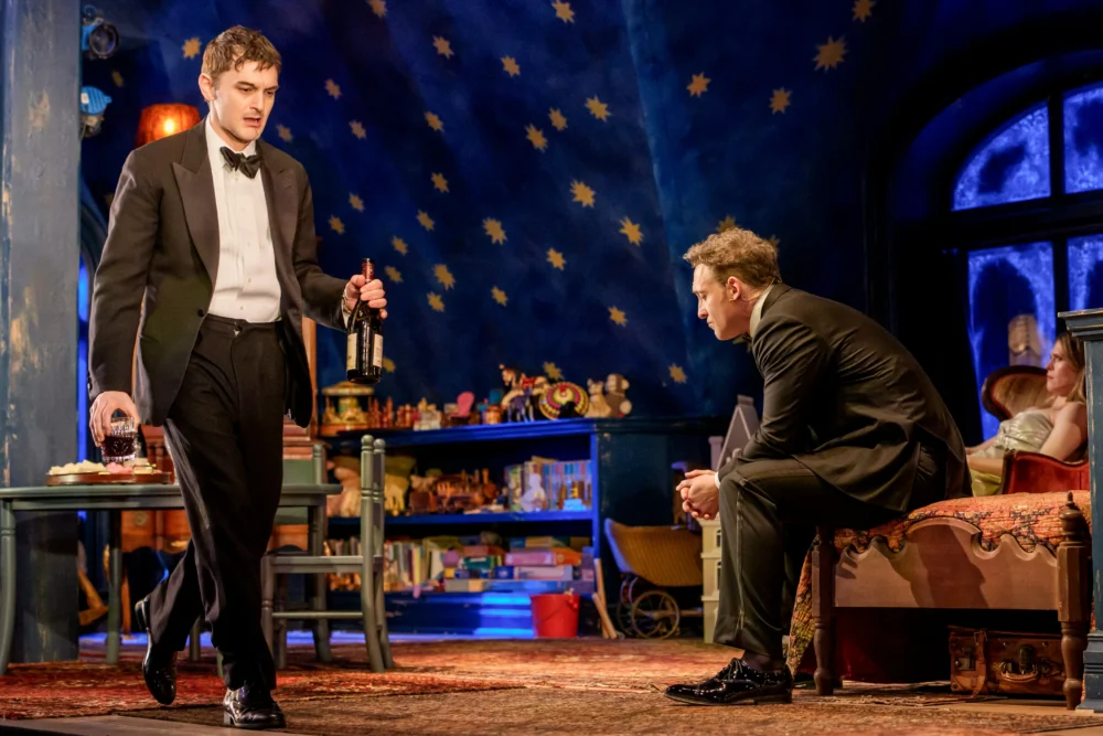 Two men in tuxedos are in a room decorated with toys and painted stars; one stands holding bottles, the other sits on a table. A woman sits in the background.